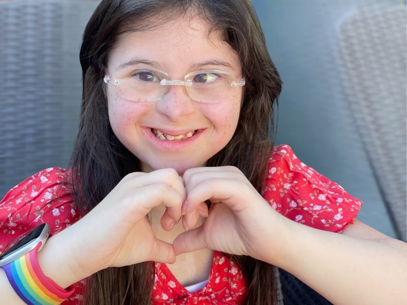 Girl smiling, making heart with hands, wearing glasses.
